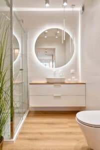 Salle de bain moderne lumineuse avec miroir rond rétroéclairé, vasque blanche sur meuble bois et blanc, douche vitrée, sol clair et plante.