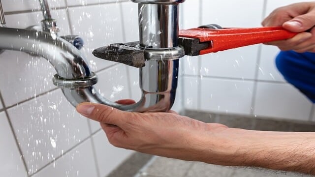 Réparation de fuite d'eau sur un tuyau de plomberie chromé sous un évier. Une main serre la clé sur le raccord, l'eau éclabousse.