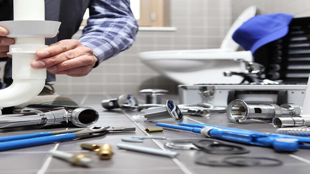 Mains d'un plombier installant un siphon d'évier en plastique blanc, outils et pièces de plomberie éparpillés sur le sol carrelé d'une salle de bain moderne.