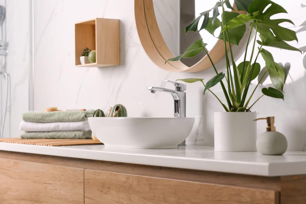 Salle de bain moderne : meuble vasque bois et design épuré Salle de bain moderne et lumineuse avec meuble vasque en bois clair, évier blanc, miroir rond et plante Monstera. Serviettes vertes empilées.
