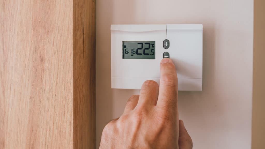 Main ajustant un thermostat digital blanc affichant 22.5 °C et 16:16, abaissant la température. Mur clair et bois en arrière-plan.